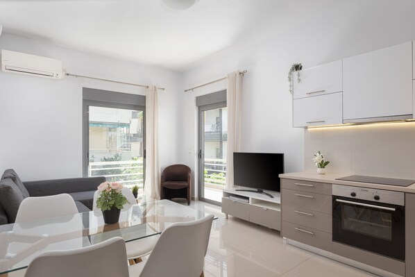 A modern living room with a glass dining table, a sofa, a TV, and a kitchen area with a stove and cabinets. - Varkiza Seaside Apartment (Vari-Voula-Vouliagmeni)