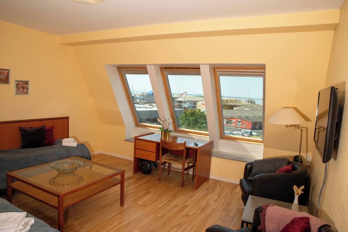 A room with a bed, a desk, a chair, a sofa, and large windows with a view of the city.