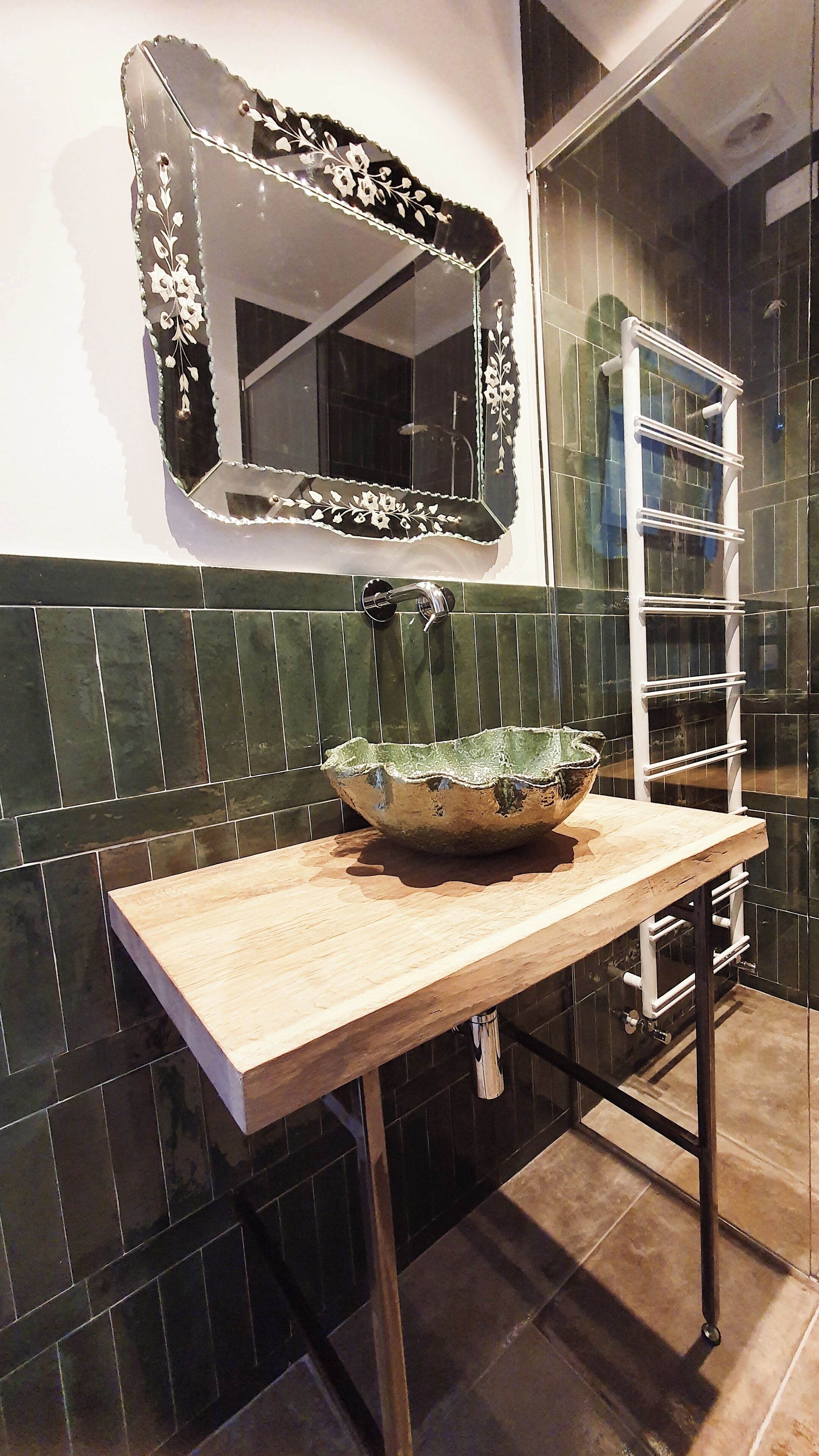 A bathroom with a wooden vanity, a green ceramic sink, a mirror with floral design, and a towel rack.