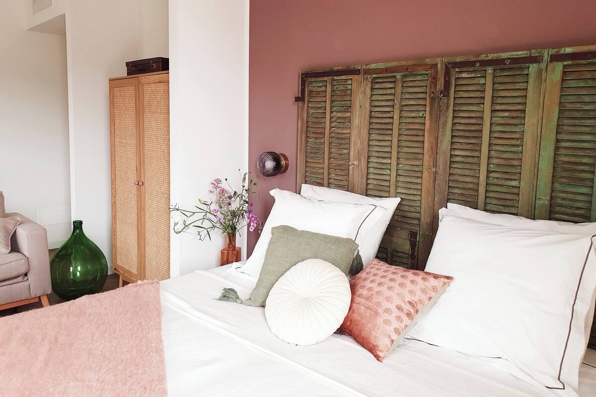 Une chambre à coucher avec un lit, des oreillers, un vase de fleurs, une bouteille verte et une armoire en bois.