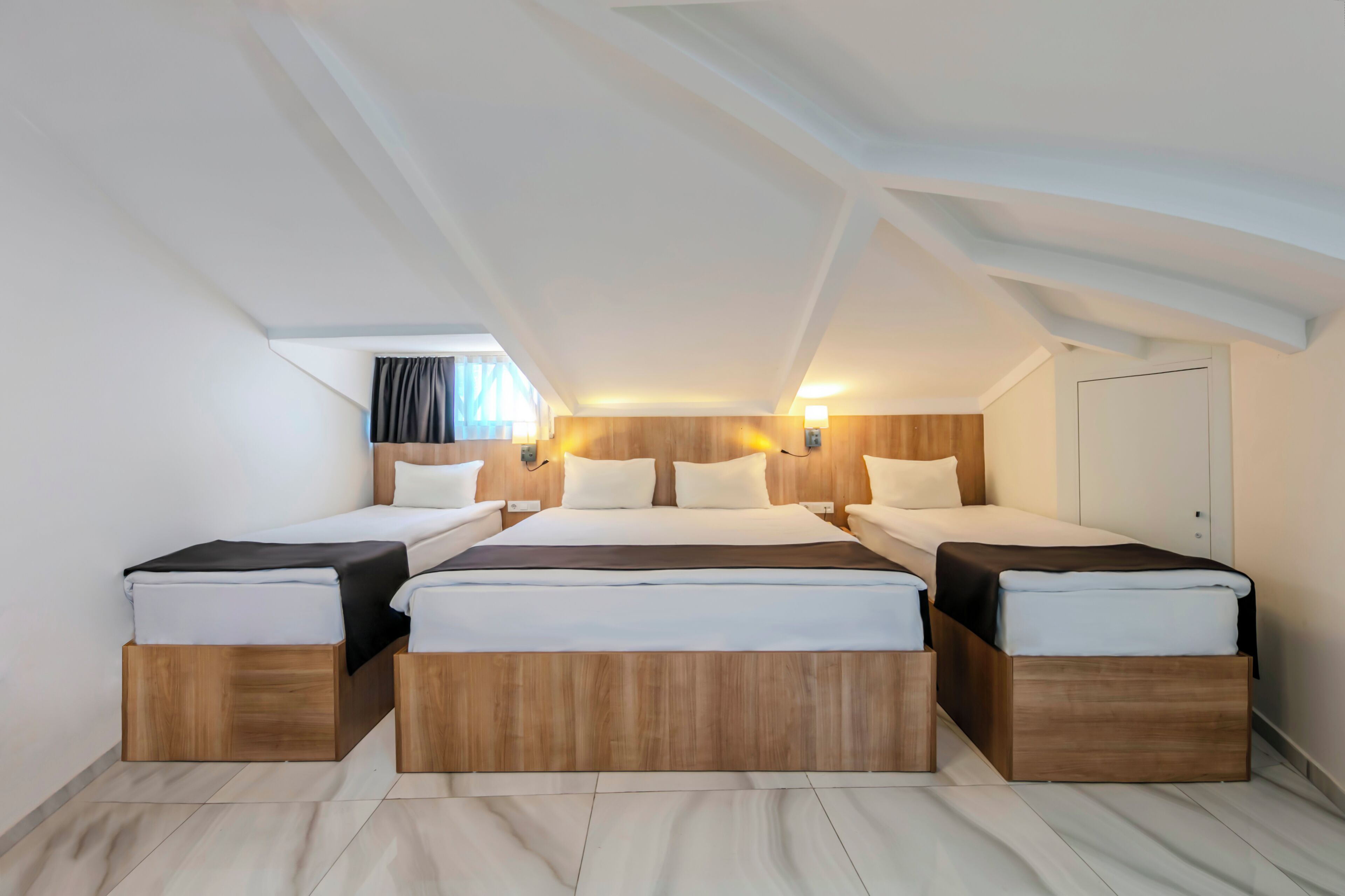 A modern hotel room with a loft-style ceiling, a large bed with a wooden headboard, two bedside tables, and a window with curtains.
