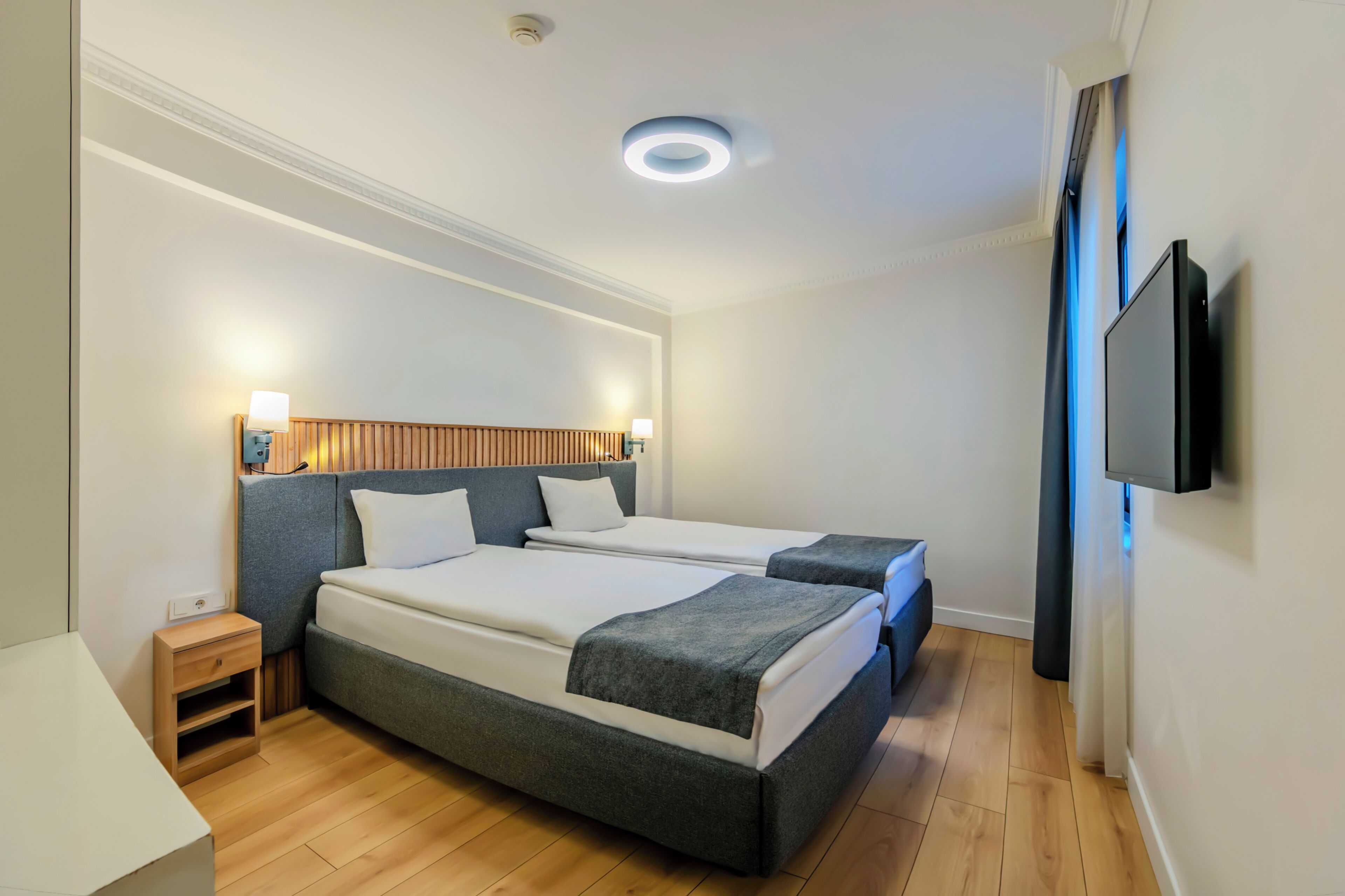 A hotel room with two beds, a wooden headboard, bedside lamps, a flat-screen TV, and a wooden floor.