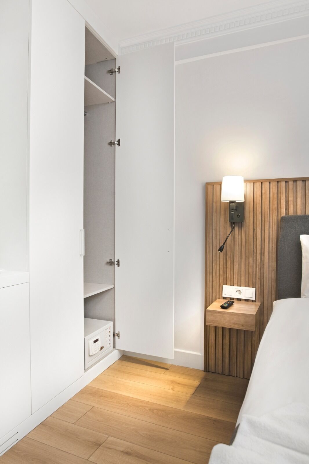 Habitación de hotel con piso de madera, una cama, una mesita de noche con lámpara y un armario con las puertas abiertas.
