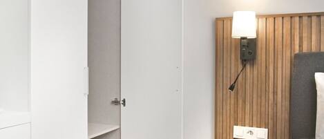 Habitación de hotel con piso de madera, una cama, una mesita de noche con lámpara y un armario con las puertas abiertas.