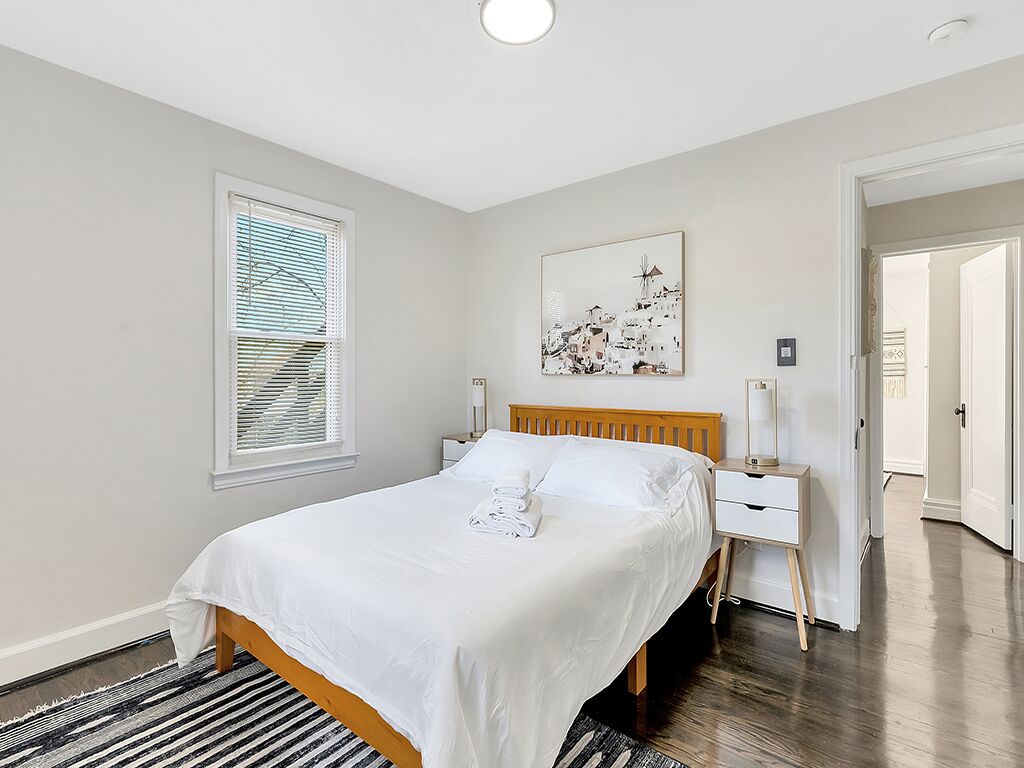 A neatly arranged bedroom with a bed, bedside tables, a window with blinds, a wall-mounted artwork, and a door leading to another room.