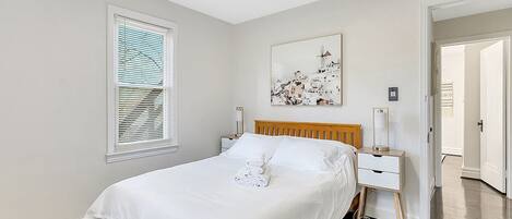 A neatly arranged bedroom with a bed, bedside tables, a window with blinds, a wall-mounted artwork, and a door leading to another room.