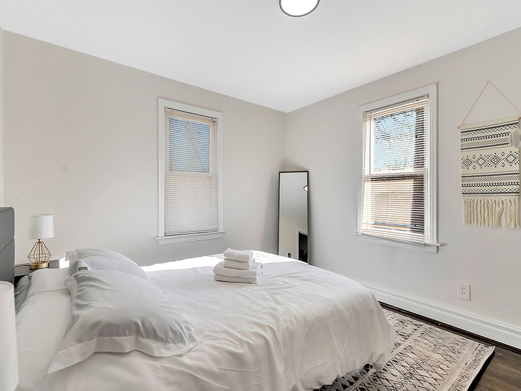 A bedroom with a large bed, two windows with blinds, a mirror, and a wall hanging.