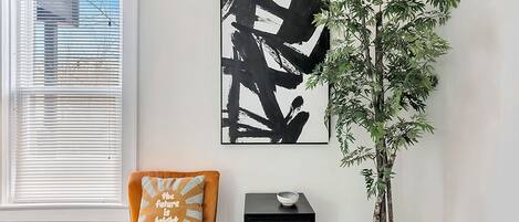 A room with a white wall, a black abstract painting, a potted plant, a black side table with a bowl, and an orange chair with a cushion.