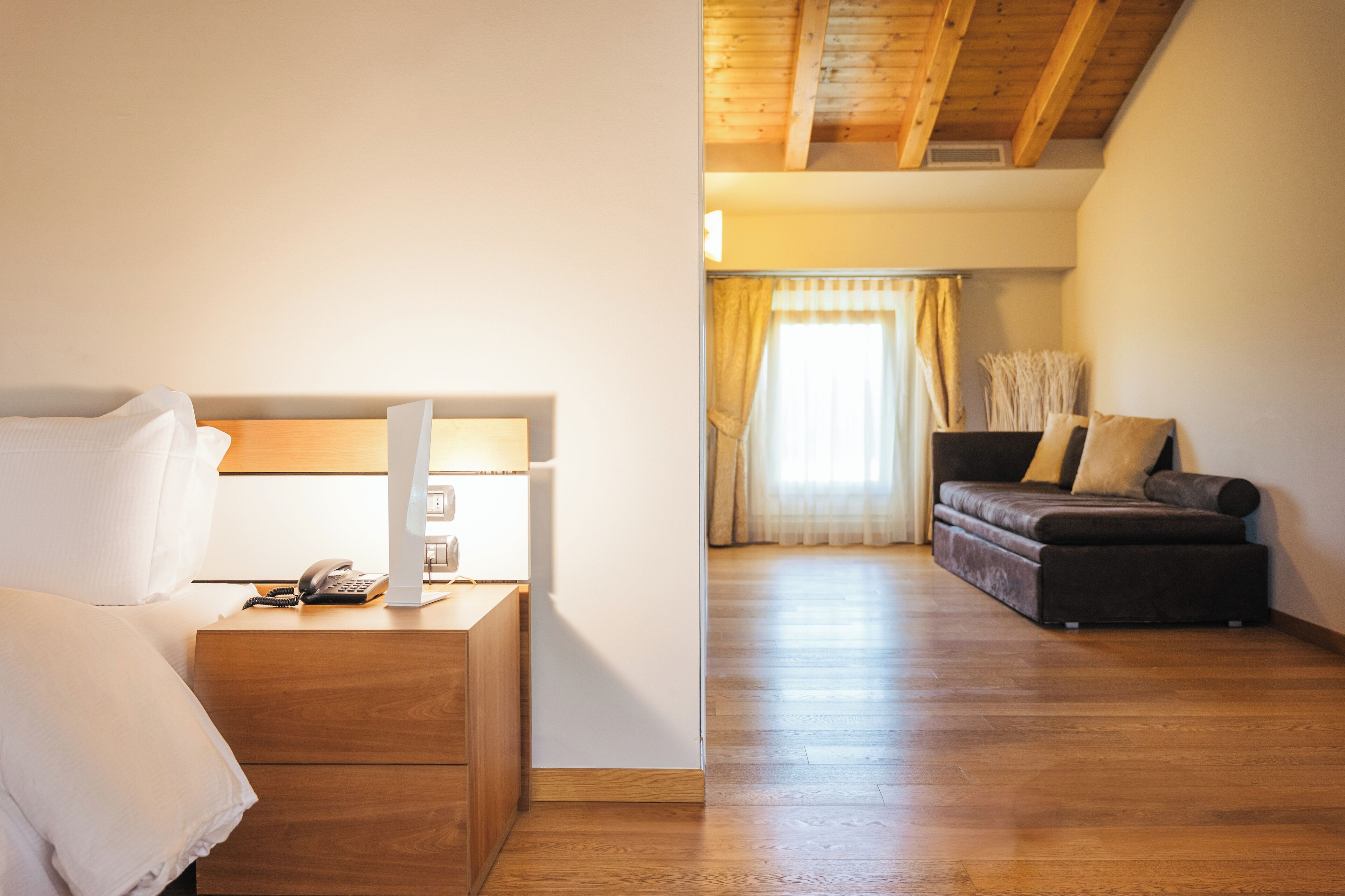 A bedroom with a wooden bed, bedside table, lamp, and a phone. There is a sofa and curtains in the adjacent living area.
