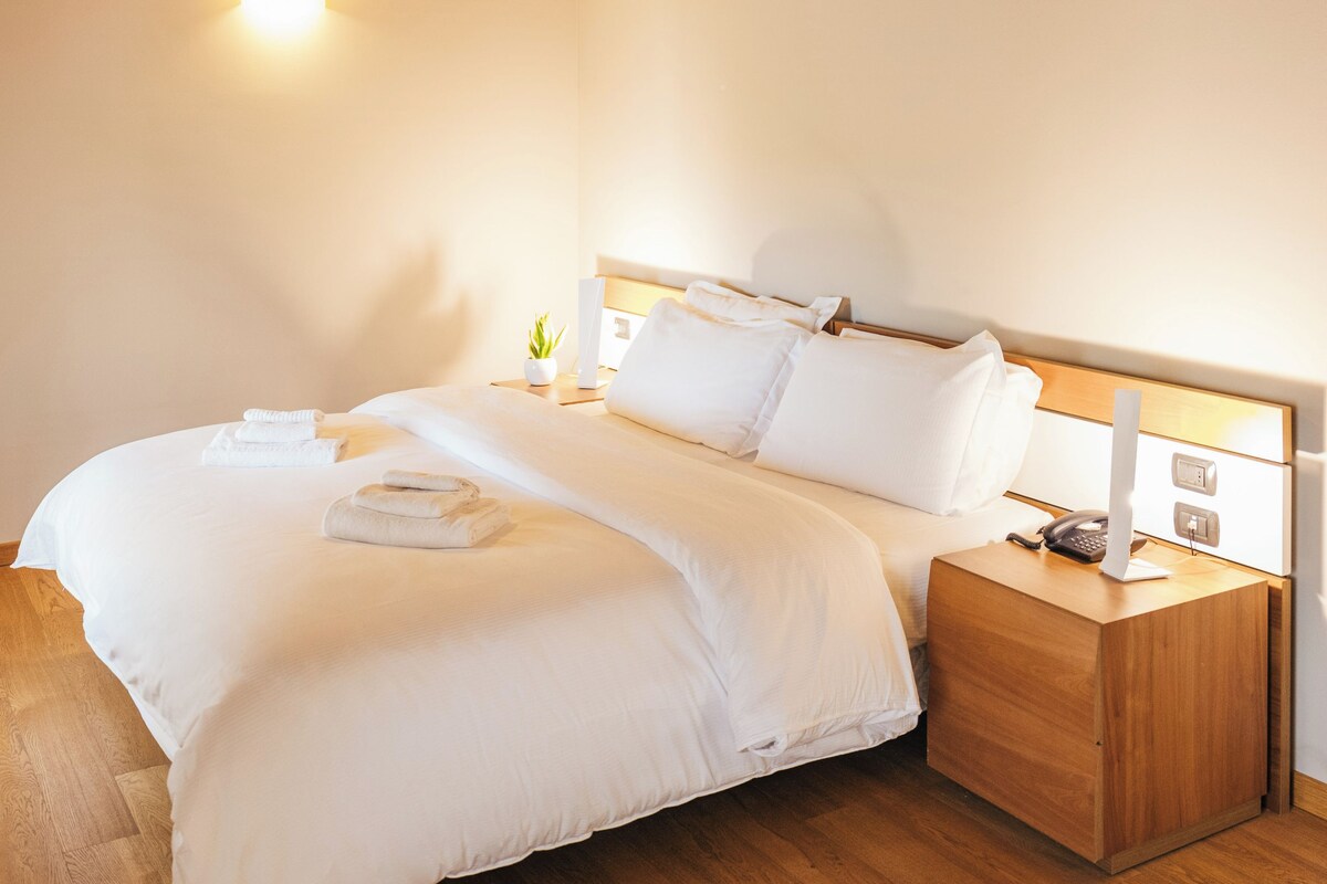 Une chambre avec un plafond en bois, un lit recouvert de draps blancs, une table de chevet avec une lampe et une petite plante.