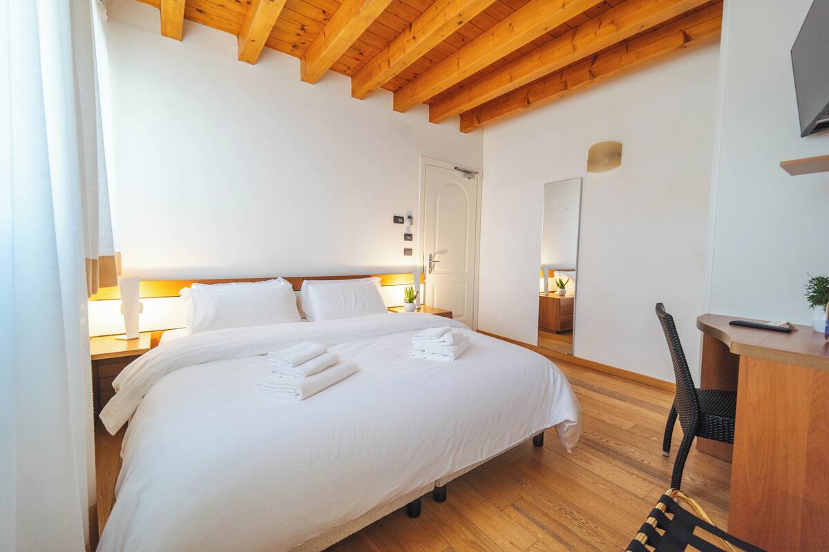 Ein Hotelzimmer mit einem großen Bett, Holzbalkendecke, einem Schreibtisch mit Stuhl und einem Spiegel an der Wand.