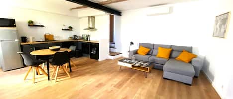 Un espacio habitable moderno con comedor, sofá gris con cojines naranja y cocina al fondo.