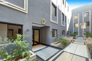 A modern building with a prominent 'D' sign, a covered entrance, and a courtyard with potted plants. - bnbmehomes-2BR nr Boulevard City-D13 (Riyadh)
