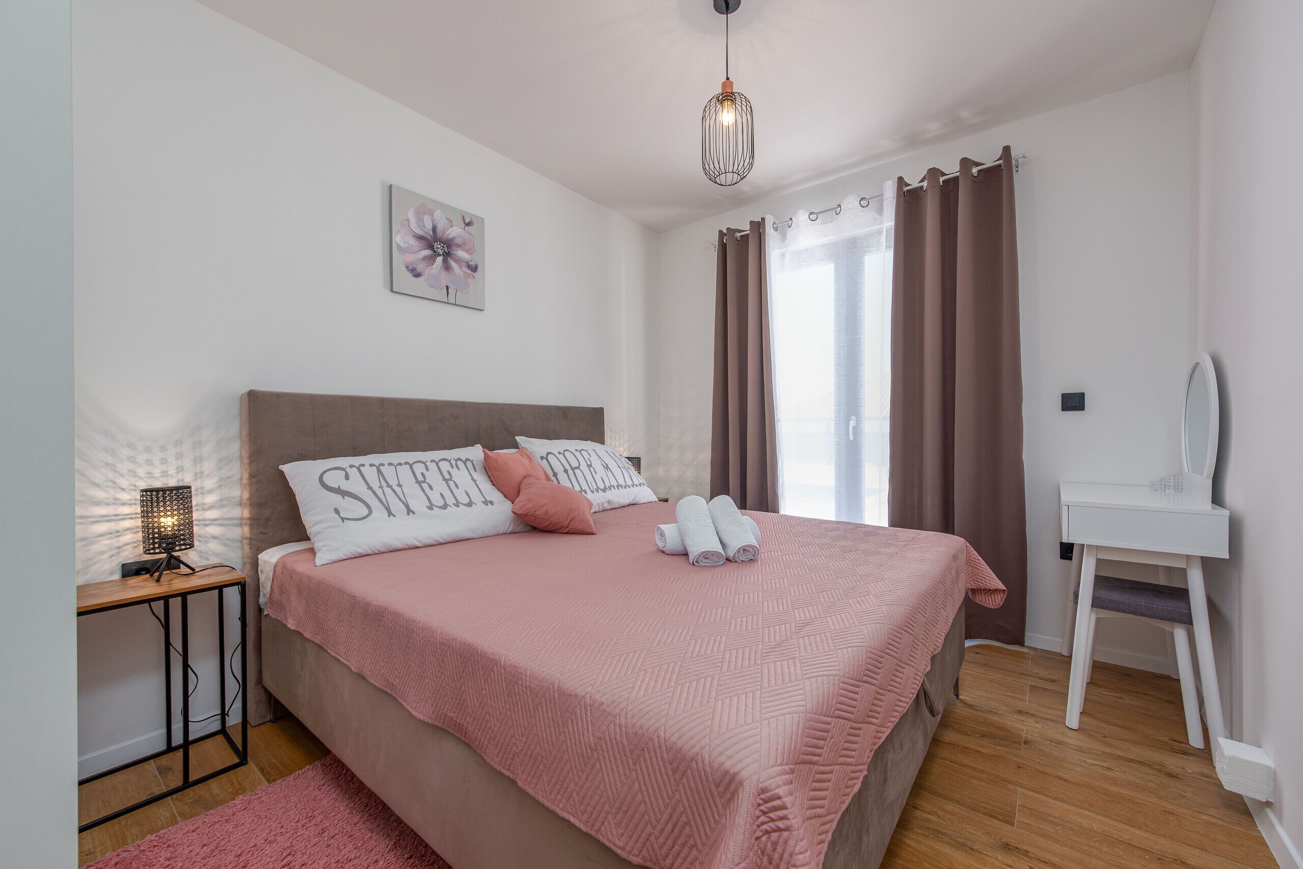 A neatly arranged bedroom with a bed, a bedside table with a lamp, a desk, and a window with curtains.