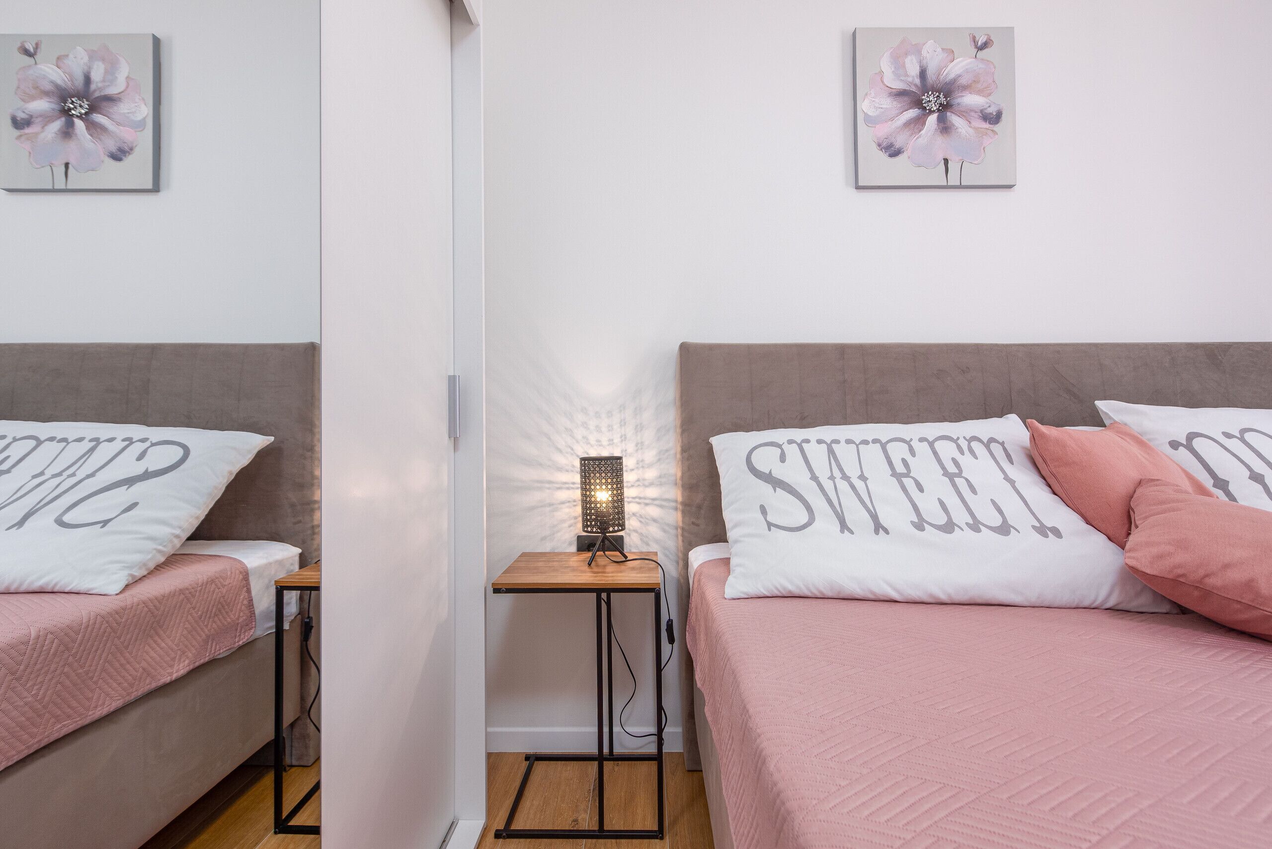 A bedroom with two beds, each with a pillow featuring the word "sweet." A small table with a lamp is between the beds.