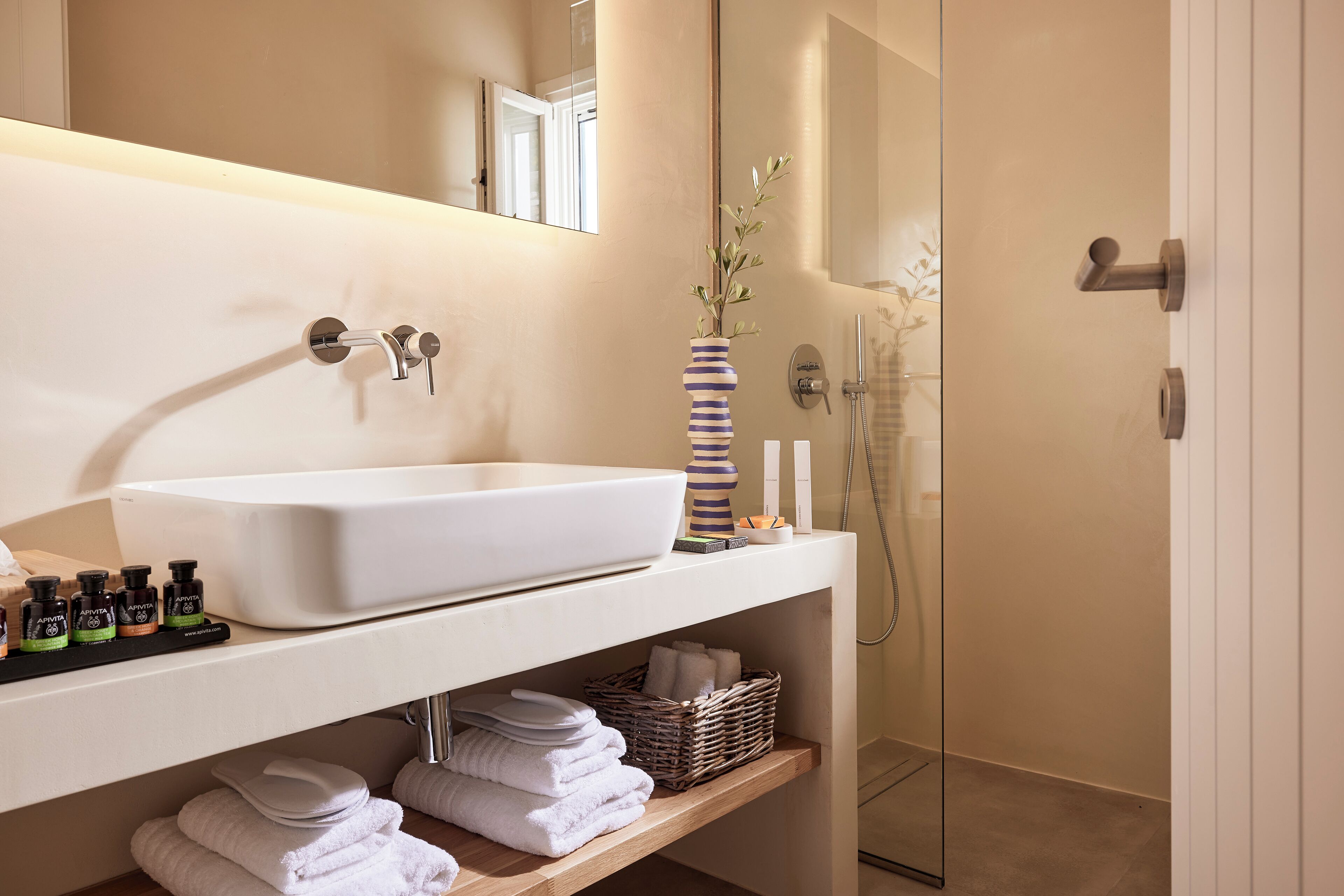 Un baño con lavamanos blanco, espejo y ducha. Hay toallas y art ículos de aseo en el mostrador.
