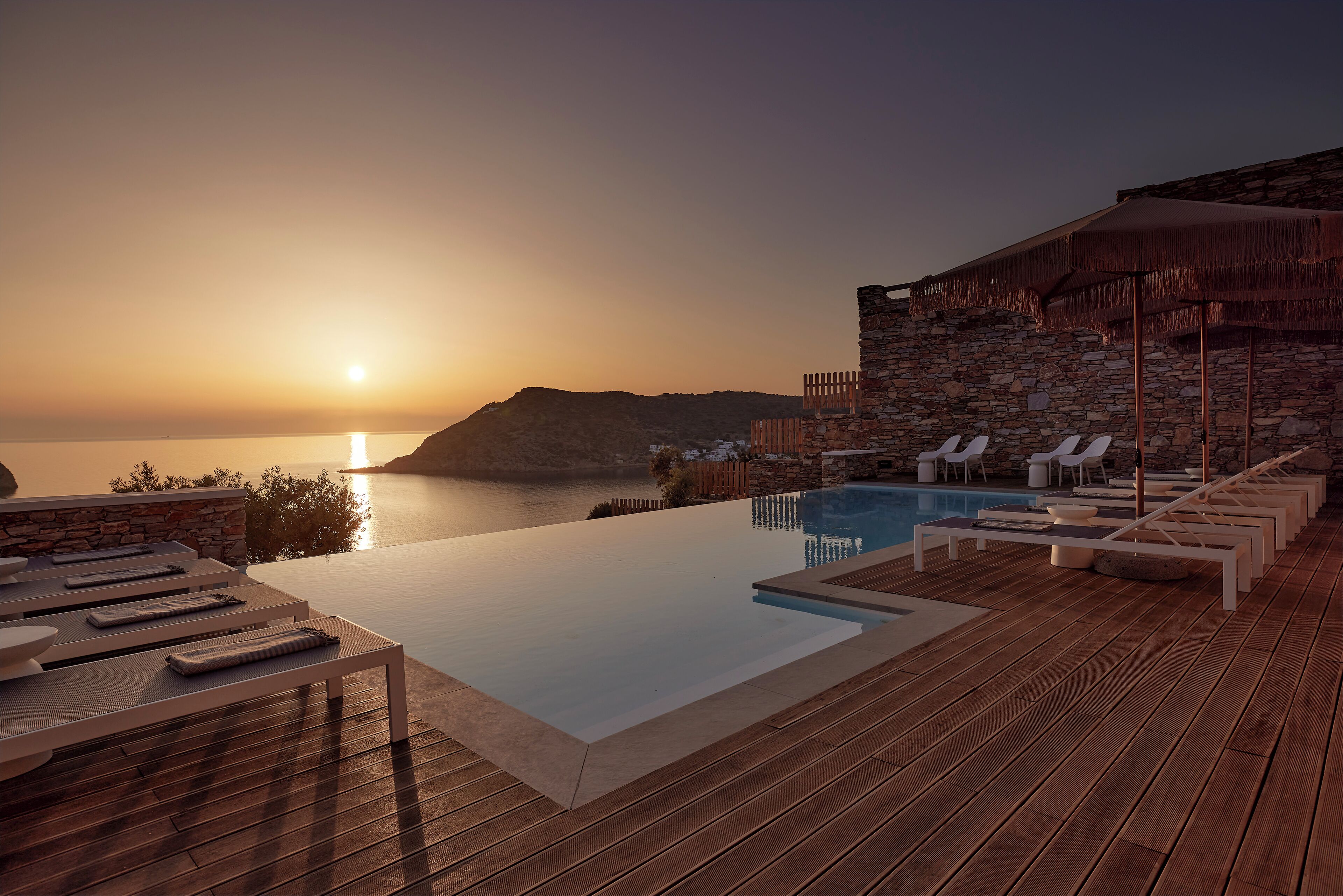 An infinity pool with lounge chairs and a stone building with a thatched roof, overlooking a body of water at sunset.