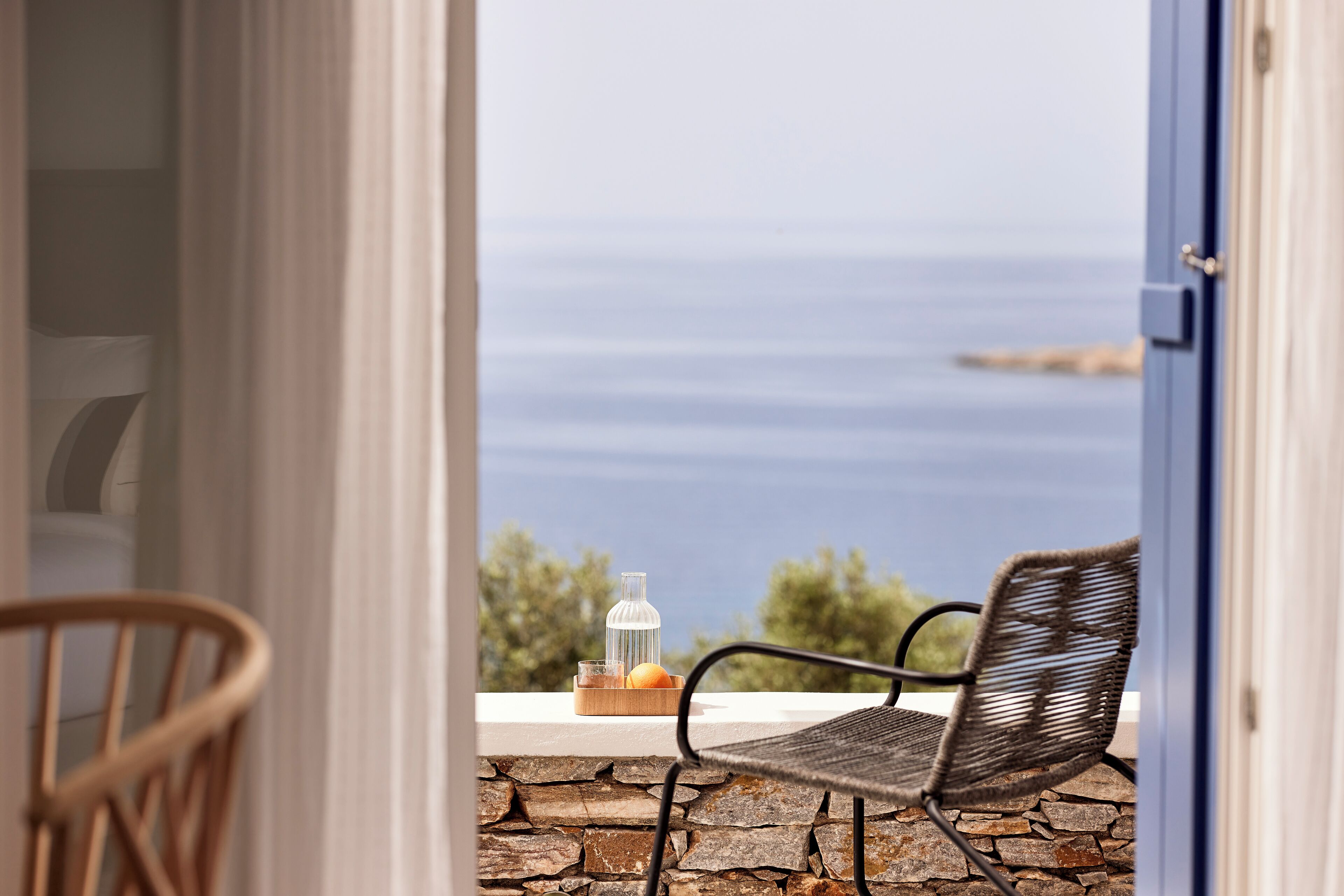 An open door leading to a balcony with a wicker chair and a table with a bottle and fruit.