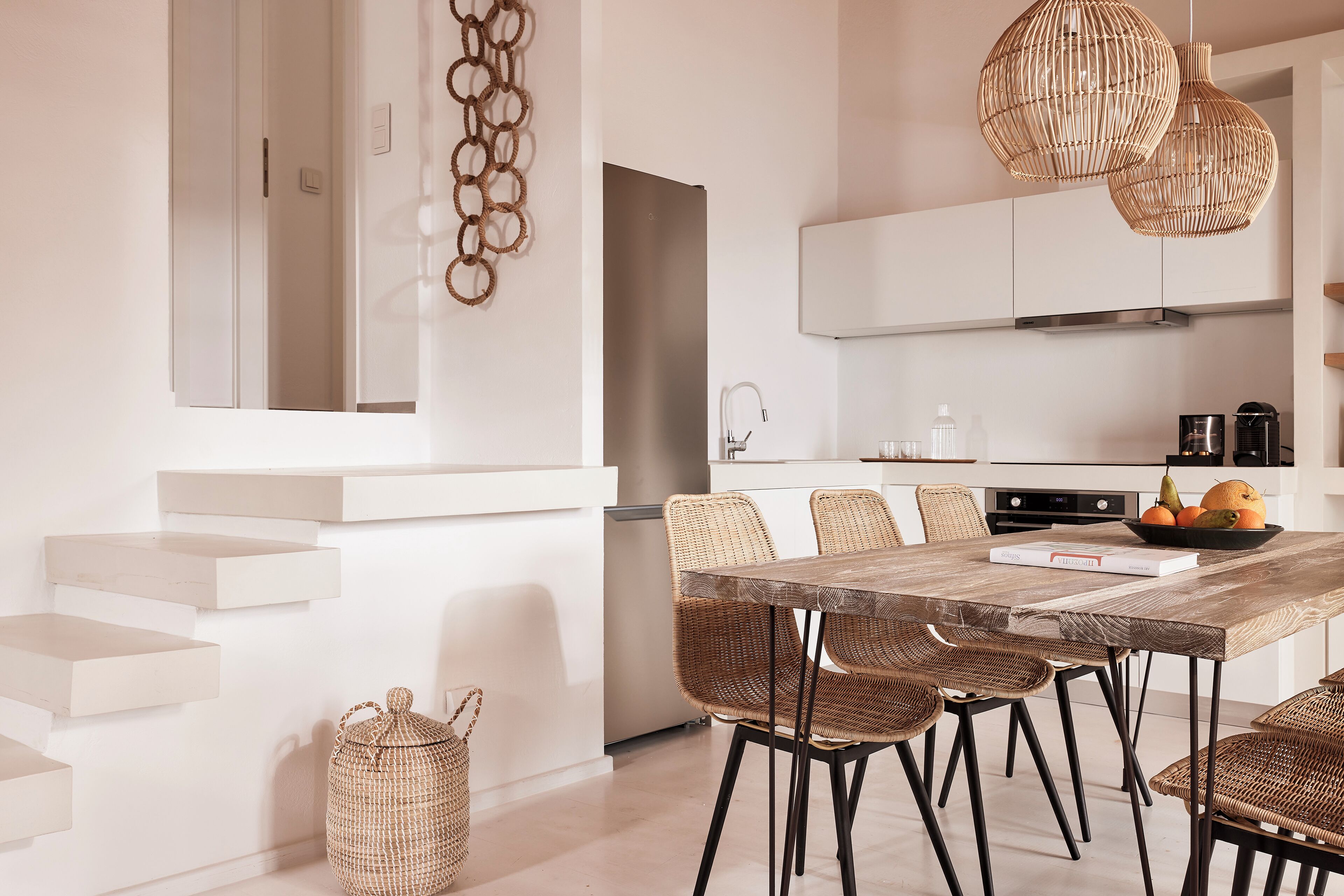 A modern kitchen with white cabinetry, a wooden dining table, wicker chairs, and a hanging light fixture.