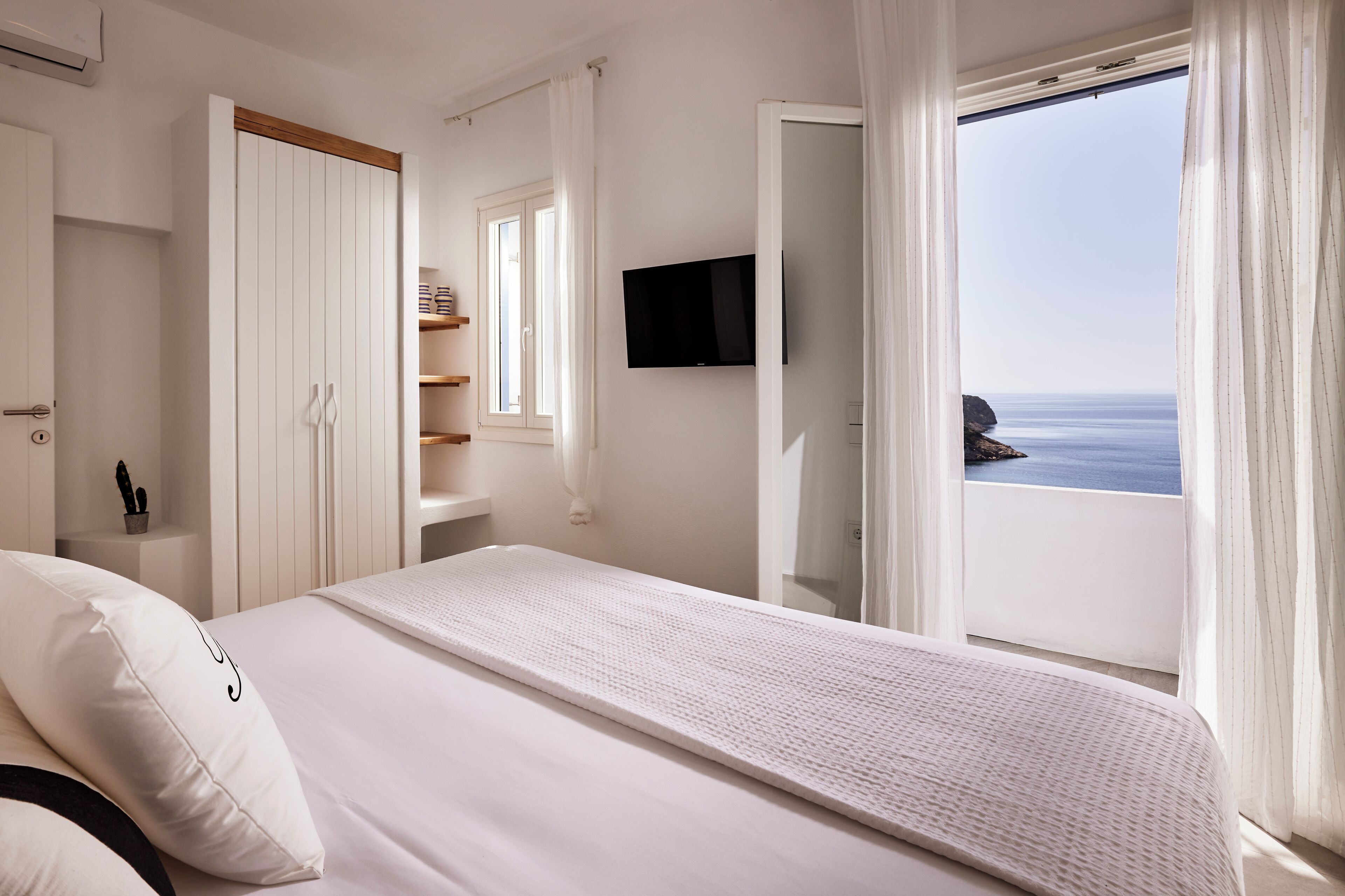 A bedroom with a bed, a TV, a window with curtains, and a view of the sea.