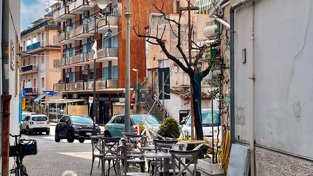 Eine schmale Straße mit parkenden Autos und einem Gassigehenden. Es gibt Gebäude mit Balkonen und eine Straßenlaterne.