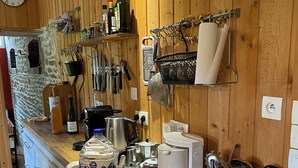 A kitchen with wooden walls, a stone wall, and a clock. It includes a refrigerator, a coffee maker, a kettle, and various kitchen utensils.