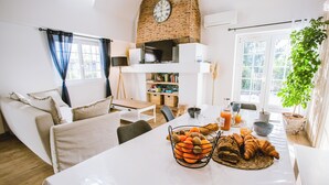 Una sala de estar luminosa con chimenea, una mesa de comedor con pan y fruta, y un sofá.