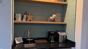 A compact kitchen with white cabinets, a black countertop, and a built-in dishwasher. There are shelves above the countertop holding decorative items and a coffee maker. - “Sound Decision” Carova Beach Canal Front Home!! 4x4 Required (Corolla)