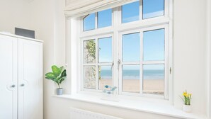 A room with a large window overlooking a beach, a white radiator, a bed with a striped bedspread, a white wardrobe, and a potted plant on the windowsill. - 1 Sunnycove, holiday property Tenby, Sleeps 4, 2 bedrooms,  bathrooms (Tenby)