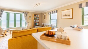 A cozy living room with a yellow sofa, a bookshelf, and a view of the sea through large windows. - 19 St Brides Bay, Holiday Apartment Broad Haven, Sleeps 6, 3 bedrooms,  bathrooms (HAVERFORDWEST)