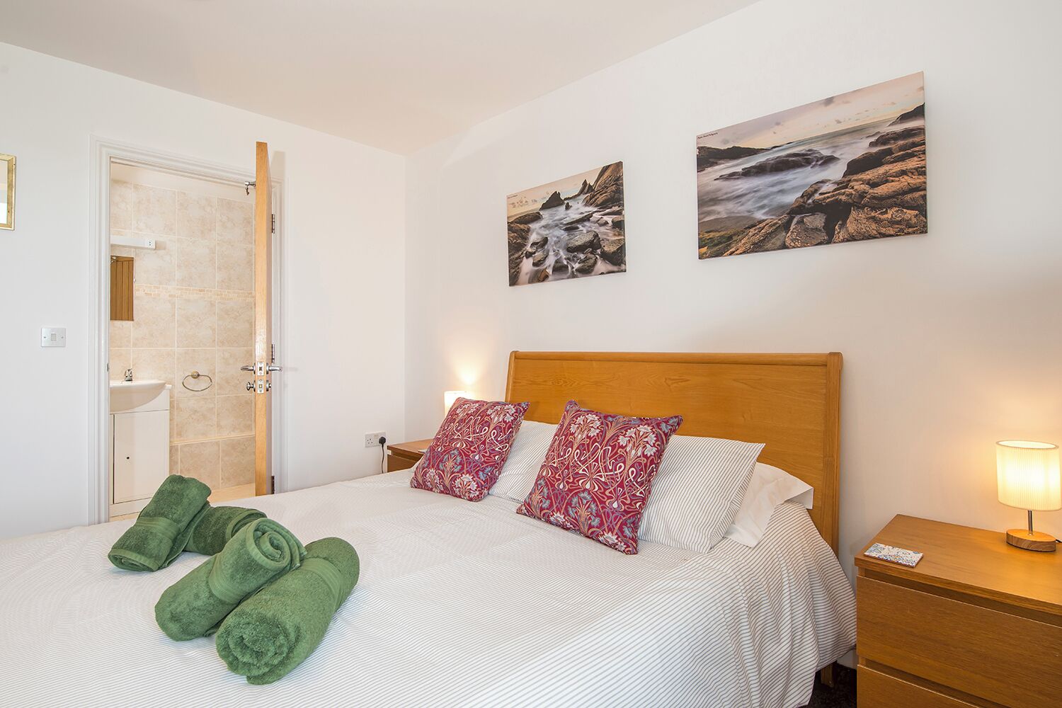 Habitación de hotel con cama, dos almohadas, una mesita de noche, lámpara, un cuadro en la pared y un baño con ducha.