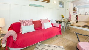 Ein gemütliches Wohnzimmer mit einem roten Sofa, karierten Kissen und einer Wanddekoration aus Holz. Im Hintergrund ist ein kleiner Essbereich mit Tisch und Stühlen zu sehen.
