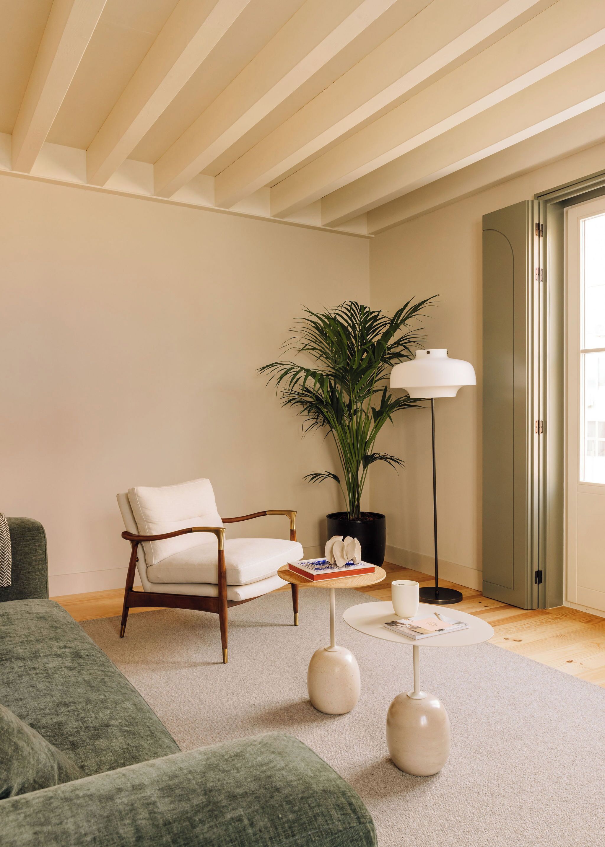Una sala de estar moderna con techo de madera, un sofá verde, un sillón blanco, una planta en maceta, una lámpara de pie y una pequeña mesa redonda con un libro y un florero.