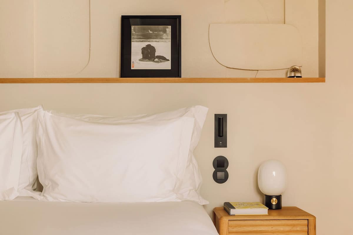 A neatly made bed with white linens, a wooden nightstand with a lamp and a book, and a framed picture on the wall.