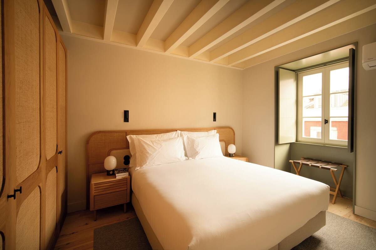 A bedroom with a large bed, wooden headboard, bedside tables with lamps, and a window with a view of buildings.