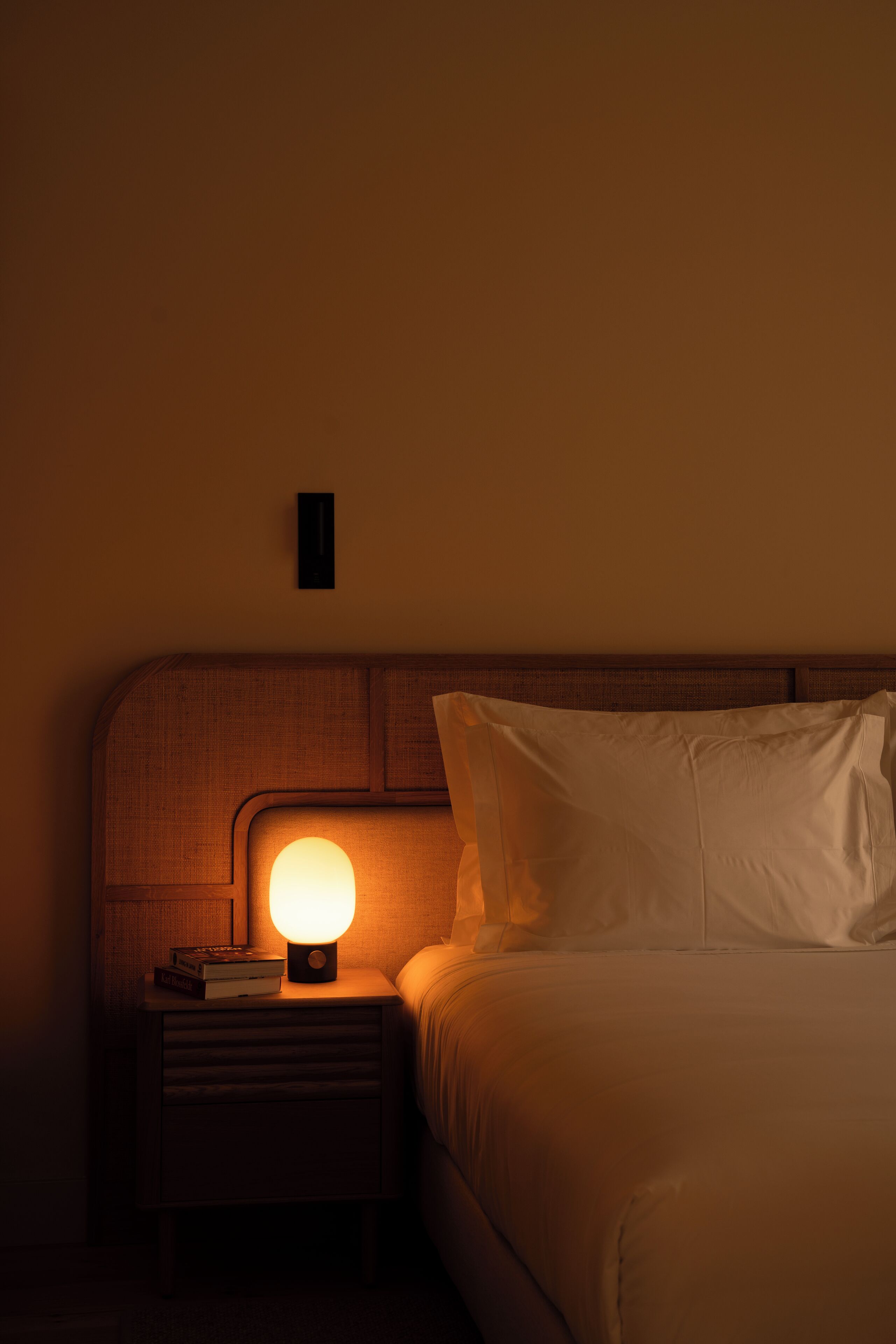 Un lit bien fait, avec du linge de lit blanc et une tête de lit en bois. On aperçoit une table de chevet avec une lampe et un livre.