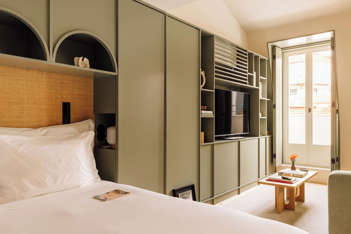A hotel room with a large bed, a TV mounted on the wall, a small table with a vase, and a view of the outside through a door.