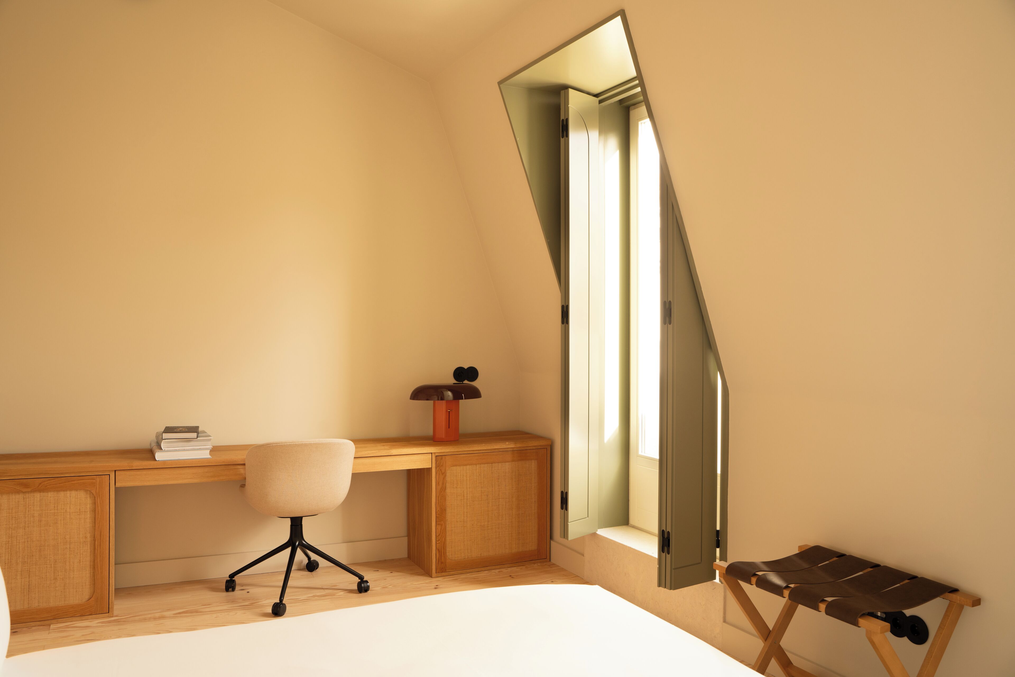 A bedroom with a sloped ceiling, a wooden desk, a chair, a bed, and a window with a view of the sky.