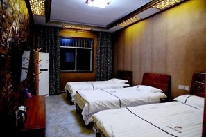 Ein Hotelzimmer mit drei Betten, einem Holztisch, einem Fenster mit Vorhängen und einer Deckenleuchte.