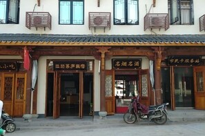 Il s’agit d’un bâtiment traditionnel doté de plusieurs entrées, certaines avec des portes en bois et d’autres avec des panneaux de verre. Des panneaux indicateurs au-dessus des portes signalent les différents services ou pièces. Une moto est garée devant le bâtiment.