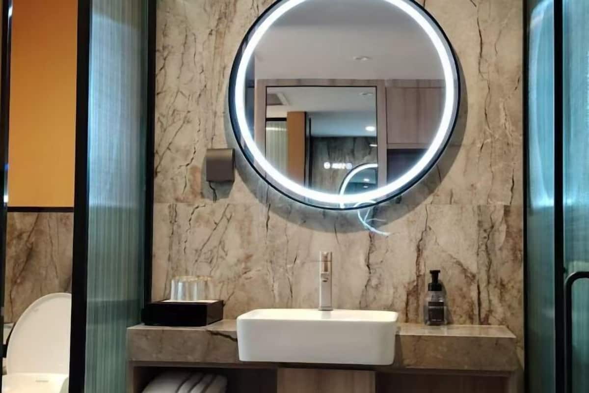 Un baño con pared de mármol, un espejo redondo con luz, un lavabo blanco y una botella de jabón.