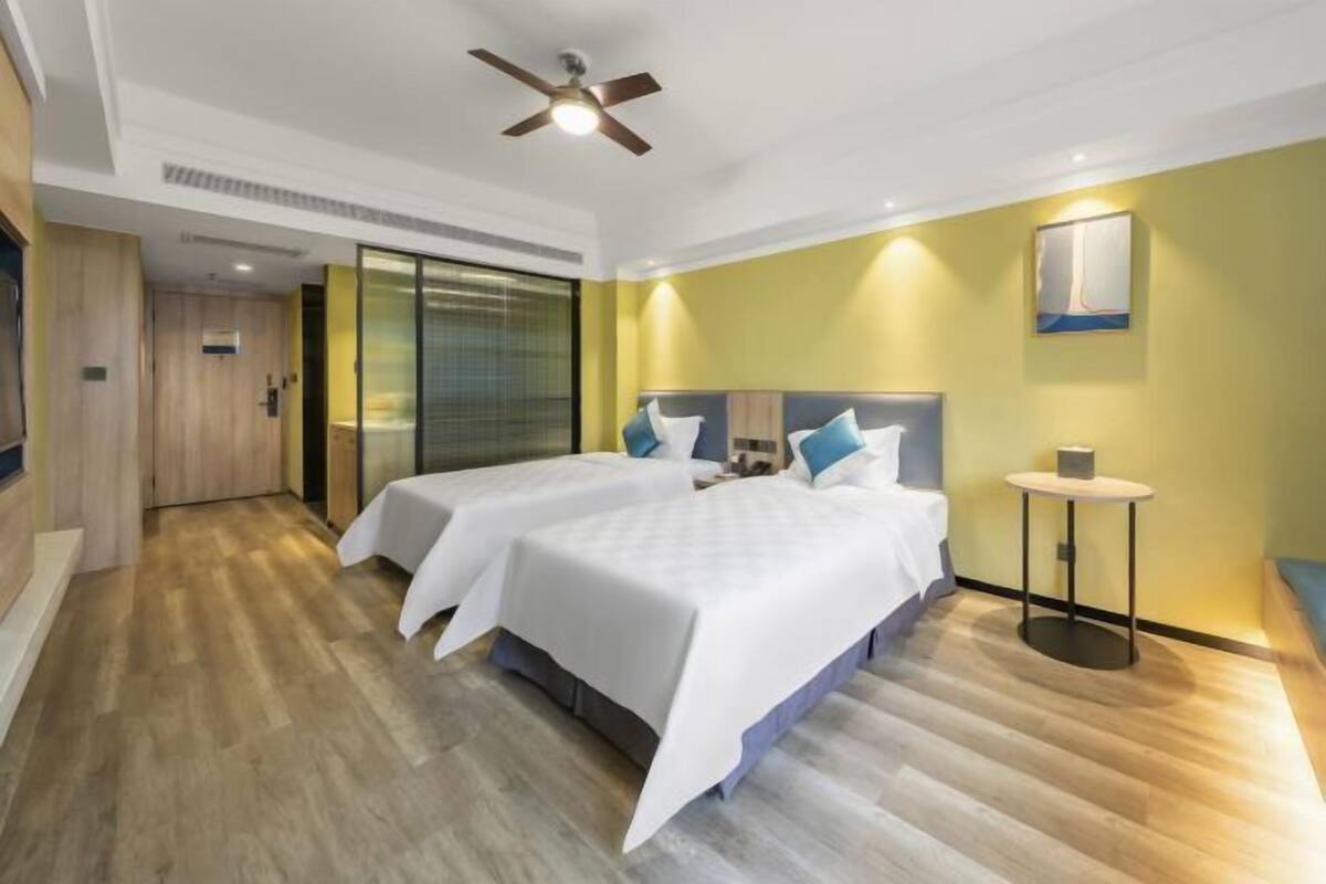Habitación de hotel con dos camas, un ventilador de techo, una mesita y una obra de arte colgada en la pared.