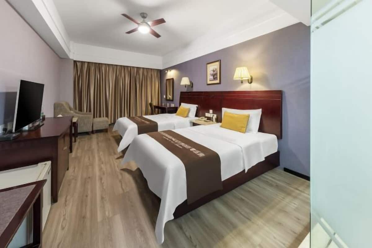 Habitación de hotel con dos camas, televisor, escritorio y ventilador de techo.