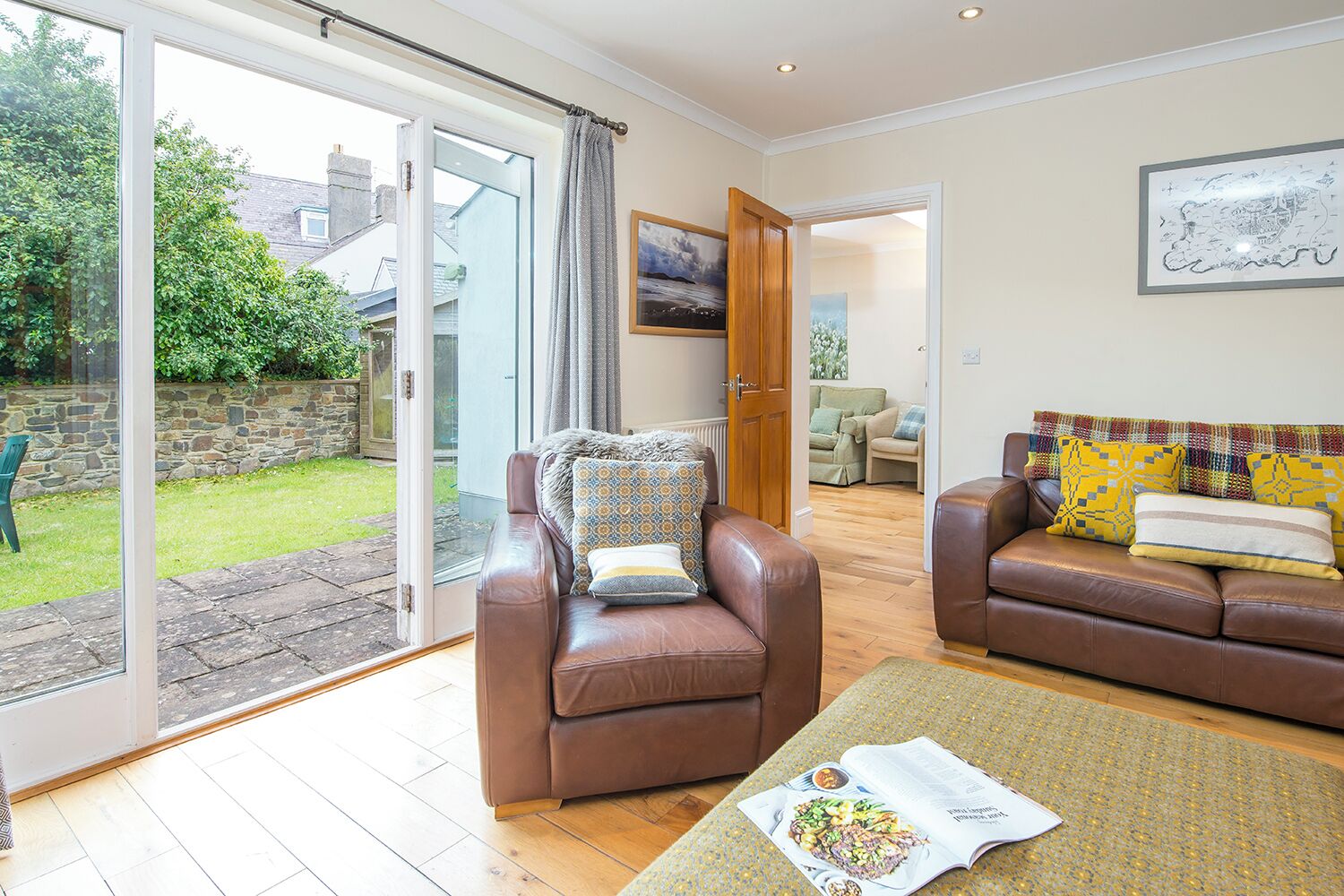 A spacious living room with a sliding glass door leading to a garden, a brown leather sofa, a matching armchair, and a dining area in the background.