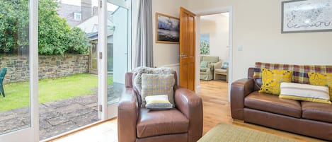 A spacious living room with a sliding glass door leading to a garden, a brown leather sofa, a matching armchair, and a dining area in the background.