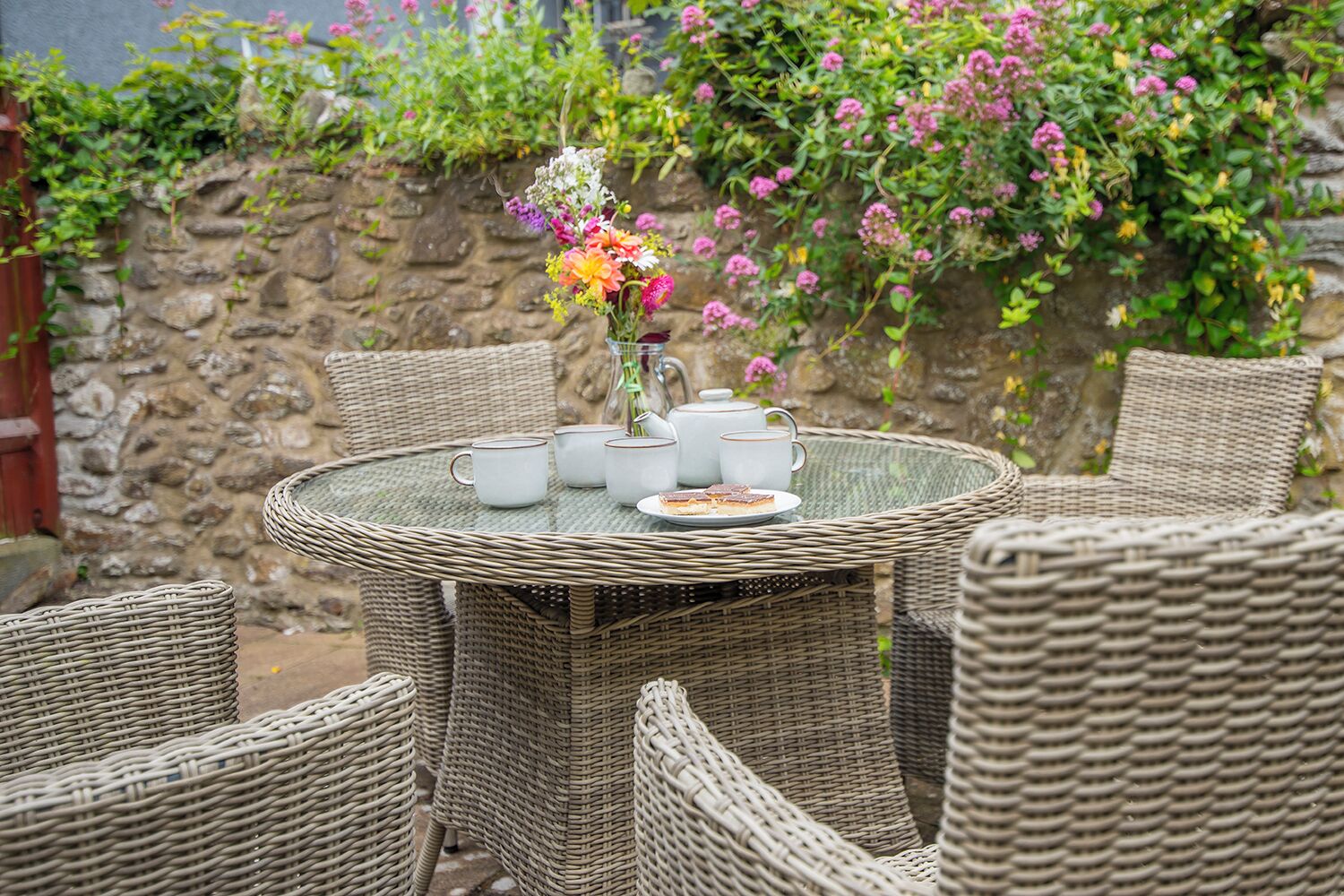 A wicker table set with white cups, a vase with flowers, and a plate with food.