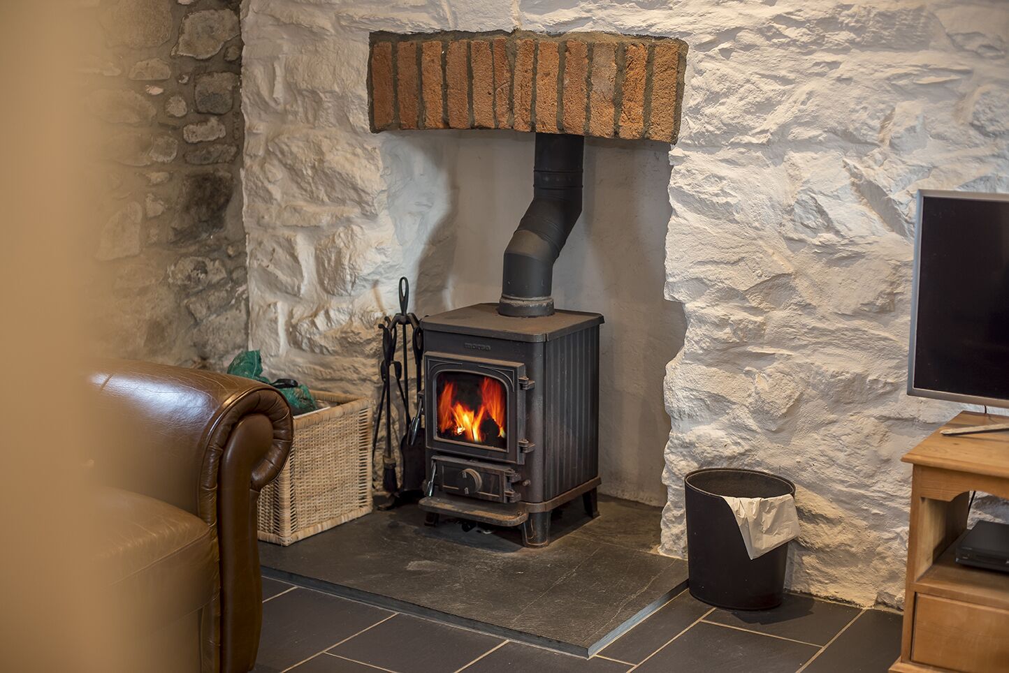 A cozy room with a stone wall, a fireplace with a visible fire, a wicker basket, and a television on a wooden stand.