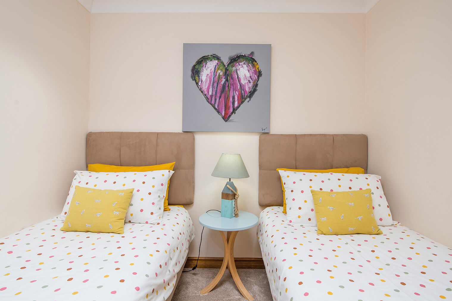 A hotel room with two beds, each with a polka dot bedspread and matching pillows. There is a small round table with a lamp between the beds. Above the table, there is a heart-shaped painting on the wall.