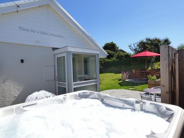 A hot tub with a glass enclosure, a patio with outdoor furniture, and a white building with a sign that reads "Time for a little relaxation."