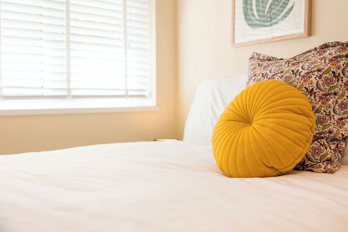 A neatly made bed with a yellow pillow and a patterned pillow, a window with blinds, and a framed artwork on the wall.